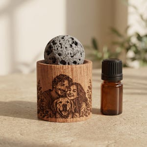 Puede incluir: Un difusor de aceites esenciales cilíndrico de madera con un retrato grabado a láser de un hombre, una mujer y un perro rodeados de follaje. Una piedra volcánica gris porosa descansa encima, junto a un frasco de vidrio ámbar con tapa negra.