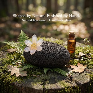 Puede incluir: Una piedra de lava negra porosa coronada con una flor de vidrio blanco translúcido con un centro amarillo, acompañada de una pequeña botella de vidrio ámbar. El texto dice: Shaped by Nature. Finished by Hand. Natural lava stone · Handmade glass.