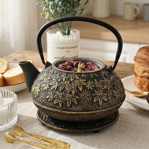 May include: A cast iron teapot with a black handle and a decorative floral pattern. The teapot is filled with dried rose buds. A glass sugar bowl, pastries, and a croissant are also on the table.