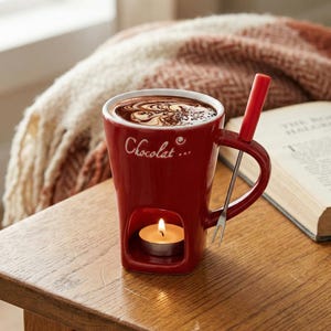 May include: A red ceramic chocolate fondue mug with the word "Chocolat" in white script. The mug has a built-in candle holder with a lit tea light. A small fork and a red stirrer are included. The mug is filled with chocolate.