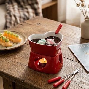 May include: A red ceramic fondue set with a heart-shaped bowl filled with melted chocolate. The set includes a small candle for heating, a fondue fork, and marshmallows. The set is on a wooden table.