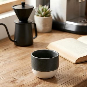 May include: A black and white ceramic coffee cup sits on a wooden surface, with a black pour-over coffee maker in the background. An open book and a small potted plant are also visible. The cup has a dark gray top and a white base.