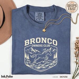 May include: A denim blue short-sleeve t-shirt featuring a cream-colored graphic of a horse jumping in front of mountains and pine trees. The text reads BRONCO OWNERS CLUB. The shirt has a Comfort Colors label at the inner neckline.