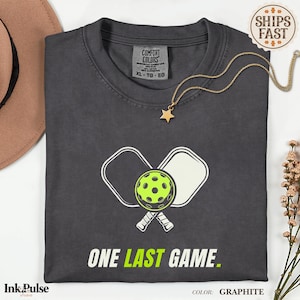 Può includere: T-shirt color grafite con grafica pickleball. Il design presenta una palla verde neon e racchette bianche, con la scritta "ONE LAST GAME". Una collana con pendente a stella dorata è appoggiata sulla maglietta.