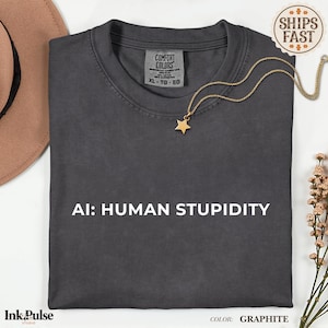 Pode incluir: Uma t-shirt de cor grafite com o texto branco AI: HUMAN STUPIDITY impresso na frente. A camisola tem um decote redondo e uma etiqueta da marca Comfort Colors no interior da gola. Um colar dourado com um pendente de estrela repousa sobre ela.