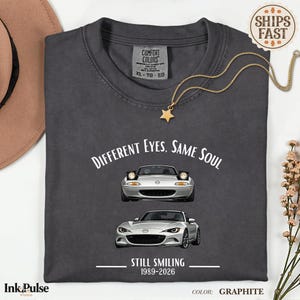 May include: A graphite-coloured t-shirt with the text "Different Eyes, Same Soul" and "Still Smiling 1989-2026". The shirt features illustrations of two silver sports cars. A gold star pendant necklace is draped on the shirt.