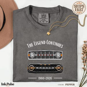 Könnte beinhalten: Ein graues Comfort Colors T-Shirt mit dem Text "THE LEGEND CONTINUES" und zwei Illustrationen eines Bronco-Grills. Das Shirt zeigt auch den Text "1966-2026" und eine goldene Sternanhänger-Halskette. Die Shirtfarbe ist Pfeffer.