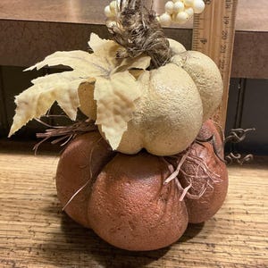 May include: A stacked pumpkin decoration featuring a textured, rust-colored bottom pumpkin and a smaller, cream-colored top pumpkin. The top is adorned with a fabric leaf, white berries, and raffia. A wooden ruler shows a height of 5 inches.
