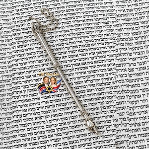 May include: A silver Yad, or pointer, with a hand-shaped tip, suspended by a chain. The pointer is used for reading a Torah scroll, with the text of the scroll in the background. A religious item.