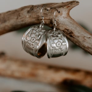 May include: A pair of silver hoop earrings with a floral design, hanging from a rustic, weathered branch. The earrings are crafted from what appears to be repurposed silverware, showcasing intricate details.