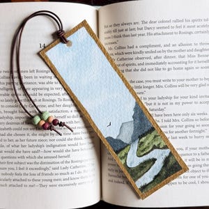 May include: A rectangular bookmark with a watercolor landscape painting featuring a winding river, green hills, and gray mountains under a light blue sky. The bookmark has a gold-painted border and a brown leather cord with two small wooden beads.