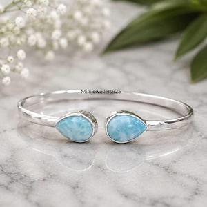 May include: A silver open cuff bracelet featuring two teardrop-shaped light blue stones at the ends. The metal band is smooth and polished. The text Minijewellers925 is visible on the inner curve of the silver band between the two stones.