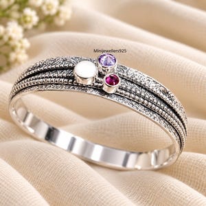 Pode incluir: Uma pulseira de prata com acabamento texturizado e oxidado, composta por três bandas sobrepostas. A pulseira é adornada com uma incrustação circular branca lisa, uma pedra preciosa redonda roxa e uma vermelha. O texto Minijewellers925 é visível.