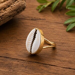 Peut inclure: Une bague dorée ornée d'un coquillage cauri blanc en son centre. Le coquillage présente une texture naturellement striée et une ouverture centrale brun foncé. L'anneau est lisse et métallique, avec une conception fendue au niveau du sertissage.
