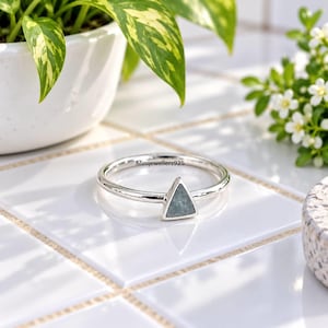 May include: A silver ring featuring a small, triangular-shaped stone in a muted blue-gray tone. The band is thin and polished, with the text Minijewellers925 clearly visible on the inner surface of the metal ring, showcasing a simple design.