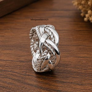 May include: A polished silver ring featuring an intricate woven design with smooth and twisted rope-like strands. The band displays a complex interlaced pattern throughout its circumference. The text Minijewellers925 is visible above the ring.