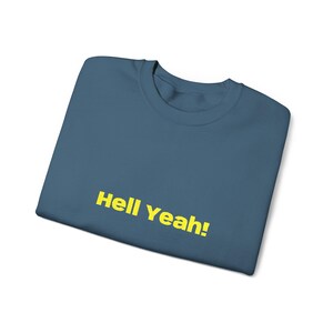 May include: A folded, teal-colored t-shirt with the phrase "Hell Yeah!" printed in yellow, centered on the front. The shirt has a crew neck and is made of a soft material.