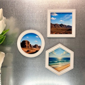 May include: Three framed landscape art pieces in white frames are displayed on a metallic surface. The art includes a round, square, and hexagonal frame, each with a different desert or beach scene. A white vase with white tulips is in the corner.