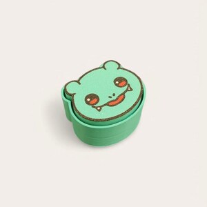 May include: A green, two-part container with a cartoon bear face on the lid. The bear face has brown outlines, red eyes, and a smiling mouth. The container is a solid, light green color.