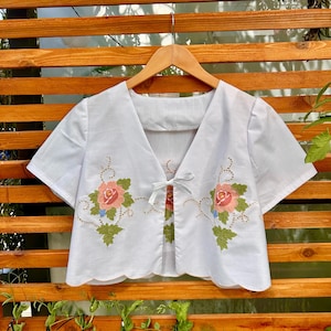 May include: White, short-sleeved top with a V-neck and a scalloped hem. The top features embroidered pink roses with green leaves and blue accents. A small bow ties the front together. The top is hanging on a wooden hanger.