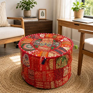 May include: A round floor pouf featuring a vibrant patchwork design in shades of red, orange, and gold. The surface is adorned with intricate embroidery, metallic thread accents, and small mirrors. It has a red piped edge and decorative tassels.