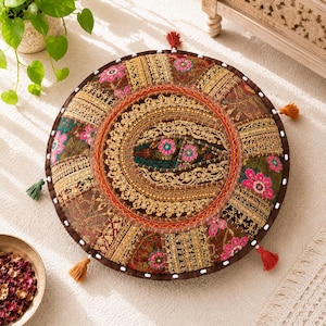 Könnte beinhalten: Runder Bodenkissen mit Patchwork-Design in Braun-, Gold- und Rosatönen. Das Kissen hat ein gesticktes zentrales Medaillon und dekorative Quasten am Rand. Das Kissen hat einen Durchmesser von etwa 50 cm.