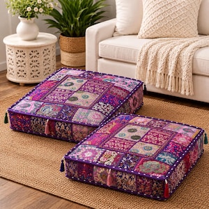 May include: Two square floor cushions featuring a vibrant patchwork design in shades of purple, pink, and magenta. Each cushion is decorated with intricate floral and geometric patterns, finished with a purple piped edge and small colorful tassels.