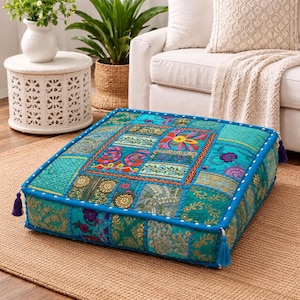 May include: A square floor cushion featuring a patchwork design in shades of turquoise and teal. The surface is composed of various fabric panels with intricate embroidery, sequins, and floral patterns, finished with blue piping and corner tassels.