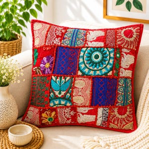 May include: A square throw pillow featuring a vibrant patchwork design with various red, blue, and green fabric panels. The surface is decorated with intricate embroidery, floral motifs, and metallic thread accents arranged in a grid pattern.