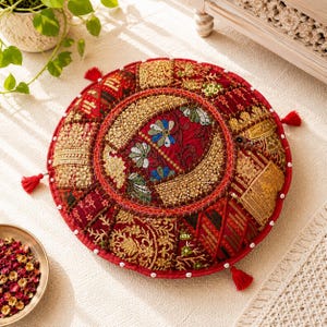 Puede incluir: Cojín de suelo redondo rojo con intrincados bordados dorados y rojos. El cojín presenta un diseño floral central, detalles de cuentas y pequeñas borlas rojas. Una pieza decorativa para interiores bohemios o de inspiración global.