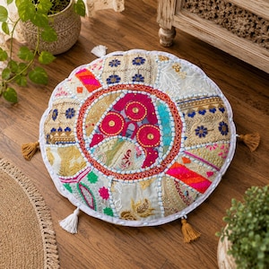 May include: A round floor cushion with a patchwork design. The cushion features a central circular pattern with pink, red, and gold accents. The outer sections are composed of various fabric patches in shades of white, blue, and orange. Tassels adorn the edges.