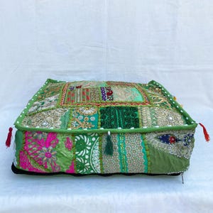 K&ouml;nnte beinhalten: Ein quadratisches Patchwork-Bodenkissen in Gr&uuml;n-, Rosa- und Goldt&ouml;nen. Das Kissen weist eine Vielzahl von Stoffmustern und -strukturen auf, mit dekorativen Quasten an den Ecken. Die Kanten sind mit einem gr&uuml;nen Rand und wei&szlig;en Perlen verziert.