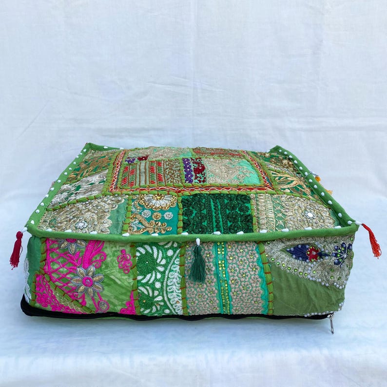 K&ouml;nnte beinhalten: Ein quadratisches Patchwork-Bodenkissen in Gr&uuml;n-, Rosa- und Goldt&ouml;nen. Das Kissen weist eine Vielzahl von Stoffmustern und -strukturen auf, mit dekorativen Quasten an den Ecken. Die Kanten sind mit einem gr&uuml;nen Rand und wei&szlig;en Perlen verziert.