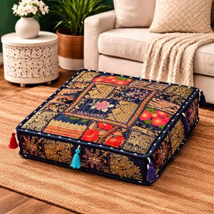 May include: Square floor cushion with a patchwork design. The cushion features a navy blue background with embroidered floral and geometric patterns in red, gold, and pink. It has tassels on the corners and white stitching around the edges.