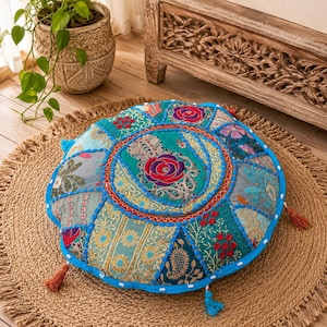 May include: Round, patchwork floor cushion with a vibrant floral design. The cushion features a mix of teal, blue, and gold fabrics, with a central floral medallion. It has a blue border and decorative tassels. Placed on a round jute rug.