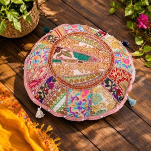 Può includere: Cuscino rotondo da pavimento con un design patchwork. Il cuscino presenta una varietà di tessuti colorati, tra cui oro, rosa, blu e verde. Ha un bordo rosa ed è adornato con piccole nappe. Il cuscino è posto su una superficie di legno.
