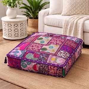 May include: Square floor cushion with a patchwork design in shades of pink, purple, and teal. The cushion has a dark purple border with white stitching and decorative tassels on the corners. The cushion is placed on a woven rug.