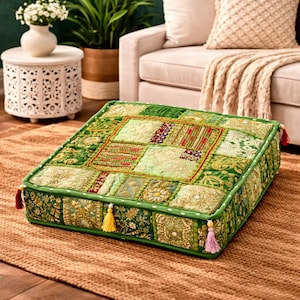 Pode incluir: Almofada de chão quadrada com um design de patchwork em tons de verde, dourado e vermelho. A almofada tem uma borda decorativa e borlas nos cantos. A almofada está colocada sobre um tapete castanho.
