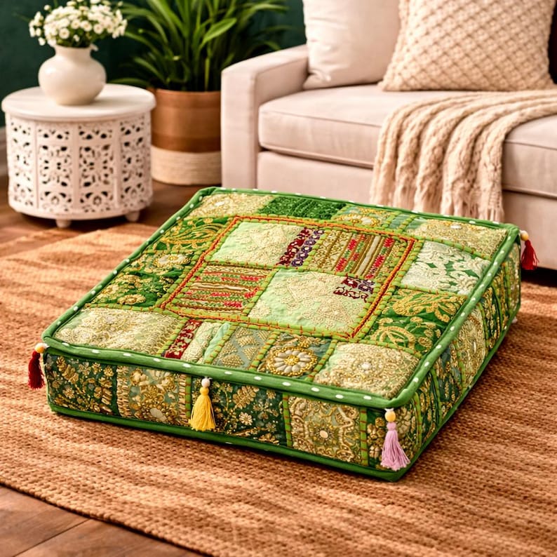 K&ouml;nnte beinhalten: Quadratisches Bodenkissen mit Patchwork-Design in Gr&uuml;n-, Gold- und Rott&ouml;nen. Das Kissen hat eine dekorative Bord&uuml;re und Quasten an den Ecken. Das Kissen liegt auf einem braunen Teppich.