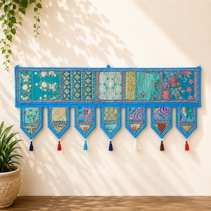 May include: A rectangular wall hanging featuring a patchwork of blue fabric panels with intricate floral and geometric embroidery. The bottom edge has seven pointed pennant-style drops, each finished with a small, colorful hanging tassel.