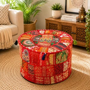 Könnte beinhalten: Ein runder Bodenpouf mit einem Patchwork-Design aus roten Stoffbahnen mit aufwendigen Goldstickereien, Pailletten und Perlenarbeiten. Der zylindrische Hocker hat eine flache Oberseite und Seiten, die mit verschiedenen Textilmustern und kleinen Quasten verziert sind.
