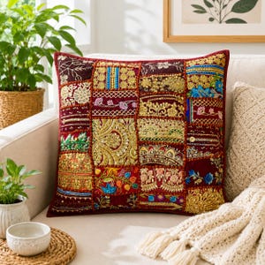 May include: A square decorative throw pillow featuring a patchwork design of deep red fabric panels. Each section is adorned with intricate gold embroidery, metallic sequins, colorful floral stitching, and beaded accents in blue, green, and pink.