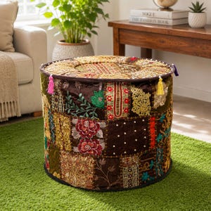 Peut inclure: Un pouf rond présentant un motif patchwork composé de divers tissus marron, dorés et rouges. La surface est ornée de broderies complexes, de motifs floraux, de fils métalliques, de petites perles blanches et de pompons colorés sur le bord.
