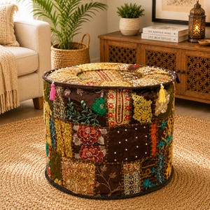 May include: A round ottoman featuring a patchwork design of various fabrics in shades of brown, gold, red, and green. The surface is decorated with intricate embroidery, floral patterns, sequins, beads, and small tassels in pink and yellow.