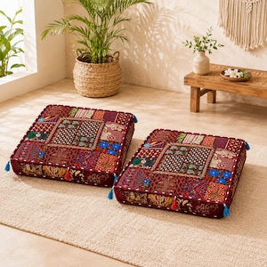 May include: Two square floor cushions featuring a patchwork design with deep red, blue, and gold fabric panels. Each cushion is decorated with intricate embroidery, small mirrors, and colorful tassels attached to the corners and sides.