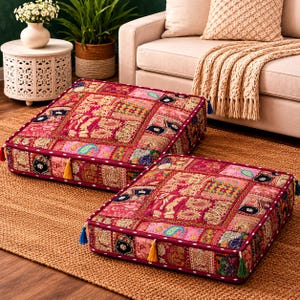 May include: Two square floor cushions featuring a patchwork design with vibrant shades of pink, red, and gold. The cushions are decorated with intricate embroidered patterns, metallic thread accents, and small colourful tassels attached to the corners.