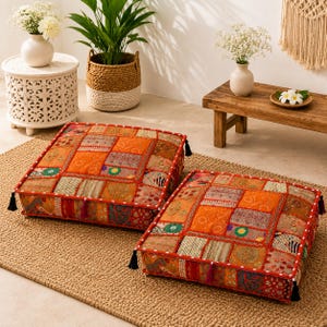 May include: Two square floor cushions featuring a patchwork design of orange, red, and gold fabric panels with intricate embroidery, beadwork, and small black tassels at each corner, finished with a white dotted trim along the edges.