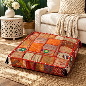 Puede incluir: Cojín de suelo cuadrado con diseño patchwork en tonos naranja, rojo y marrón. El cojín tiene un borde rojo con costuras blancas y borlas negras en las esquinas. Se encuentra sobre una alfombra de yute tejida.