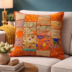 Könnte beinhalten: Quadratisches orangefarbenes Patchwork-Kissen mit bestickten und gemusterten Quadraten. Das Kissen hat einen durchgehenden orangefarbenen Rand und liegt auf einem neutralen Sofa. Ein dekoratives Wohnaccessoire.