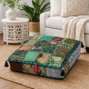 Puede incluir: Cojín de suelo cuadrado con un diseño de patchwork en tonos verdes, turquesa y dorado. El cojín tiene un borde verde oscuro con costuras blancas y borlas decorativas en cada esquina. El cojín está colocado sobre una alfombra de yute tejida.